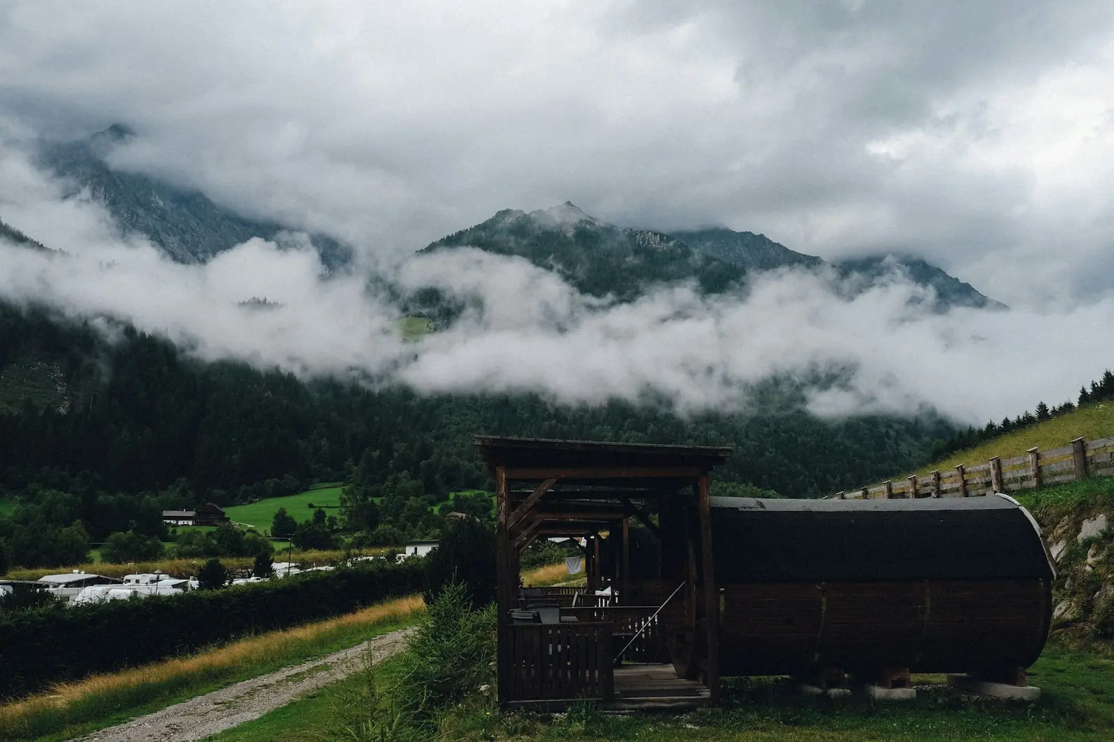 A barrel sauna and a wooden shelter in a green valley under misty mountains